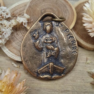 Könnte beinhalten: Ein bronzener Anhänger mit einer Darstellung eines Heiligen, der ein Boot hält. Der Anhänger trägt die Inschrift "St. Pierre" auf der Seite.