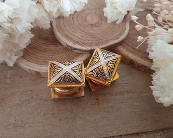 May include: Two gold-toned square cufflinks with ornate silver and black detailing. The cufflinks feature a cross design and are set against a wooden background with white floral accents. Each cufflink measures approximately 0.75 inches square.