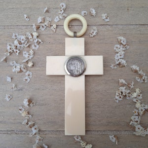 Op de afbeelding: Een wit houten kruis met een zilveren medaillon met een profiel van een persoon. Het kruis hangt aan een witte ring en is omringd door witte bloemblaadjes op een lichtbruine houten achtergrond.