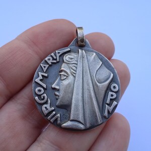 May include: A circular silver-toned metal pendant featuring a relief profile of a figure wearing a head covering. The text VIRGO MARIA is embossed along the left edge, with OPN on the right. A small metal loop is attached to the top for suspension.