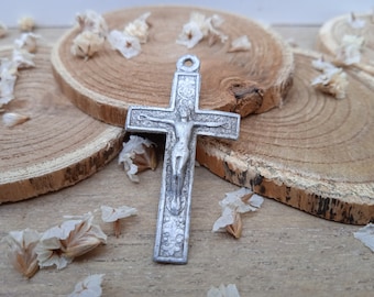 Peut inclure: Un pendentif en métal argenté en forme de croix avec un design détaillé. La croix représente une figure de Jésus-Christ avec une couronne d'épines. Le pendentif est présenté sur une surface en bois avec des pétales de fleurs blanches.