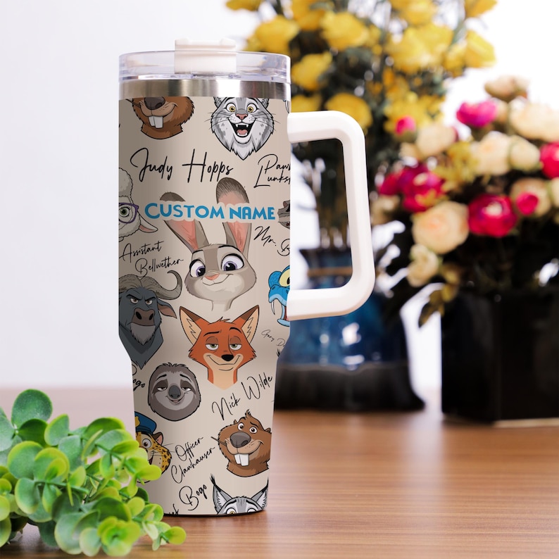 Puede incluir: Un vaso de acero inoxidable con ilustraciones de personajes de dibujos animados y el texto "Judy Hopps" y "Custom Name". El vaso tiene un asa y tapa blancas, con un fondo beige.