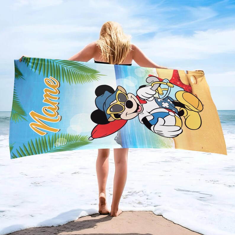 以下が含まれることがあります： サングラスと帽子をかけた漫画のネズミがビーチのシーンを背景にしたビーチタオル。タオルには筆記体で「Mame」という名前が入っています。左側にはヤシの葉があります。海に立っている人がタオルを持っています。