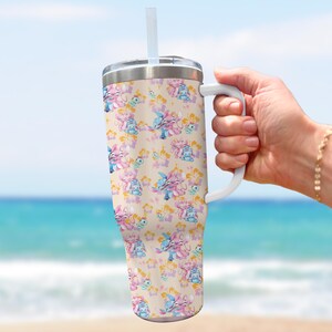 Puede incluir: Un vaso grande y aislado con asa y pajita blancas. El vaso presenta un patr&oacute;n repetido de personajes de dibujos animados en rosa y azul sobre un fondo beige, con elementos florales.
