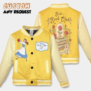 Puede incluir: Chaqueta universitaria amarilla con mangas color crema. La parte delantera presenta un dibujo de una mujer con un vestido azul y el texto "Belle's Book Shop". La parte trasera muestra "Belle's Book Club" con una rosa en una cúpula de cristal, libros y las palabras "As Time" y "1991".