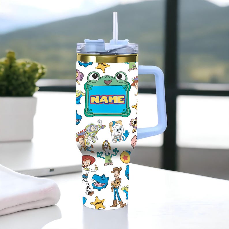 May include: A white stainless steel tumbler with a light blue handle, a clear lid, and a straw. The exterior features a repeating pattern of cartoon characters and the word NAME inside a green frog-shaped frame, including figures like a cowboy, a space ranger, and a cowgirl.