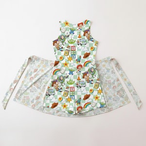 Puede incluir: Un delantal sin mangas con un estampado de cuadros verde claro y blanco, con personajes y objetos de dibujos animados coloridos. El delantal tiene lazos largos y un estampado a juego en los lazos y la falda del delantal.