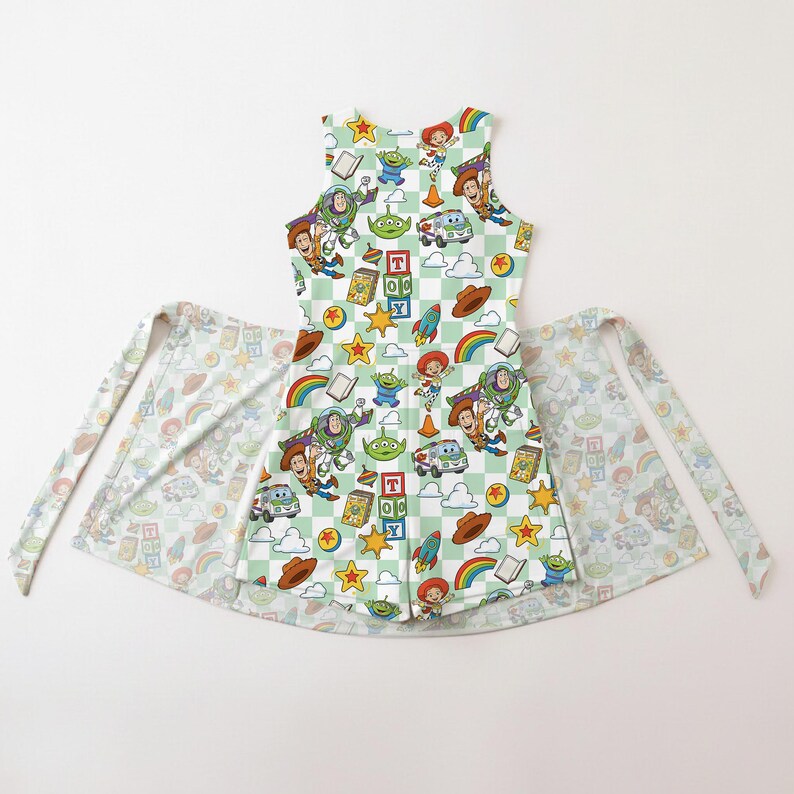 Puede incluir: Un delantal sin mangas con un estampado de cuadros verde claro y blanco, con personajes y objetos de dibujos animados coloridos. El delantal tiene lazos largos y un estampado a juego en los lazos y la falda del delantal.