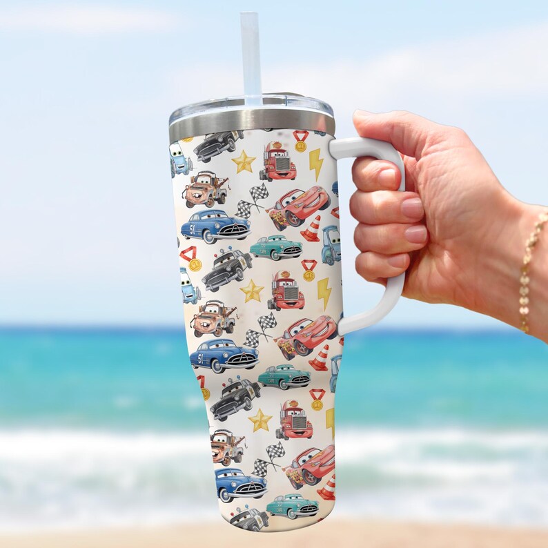 May include: A white tumbler with a handle and a straw, featuring a colorful pattern of cartoon cars, stars, and racing flags. The design includes red, blue, and green cars, along with lightning bolts and medals. The tumbler is held against a blurred beach background.