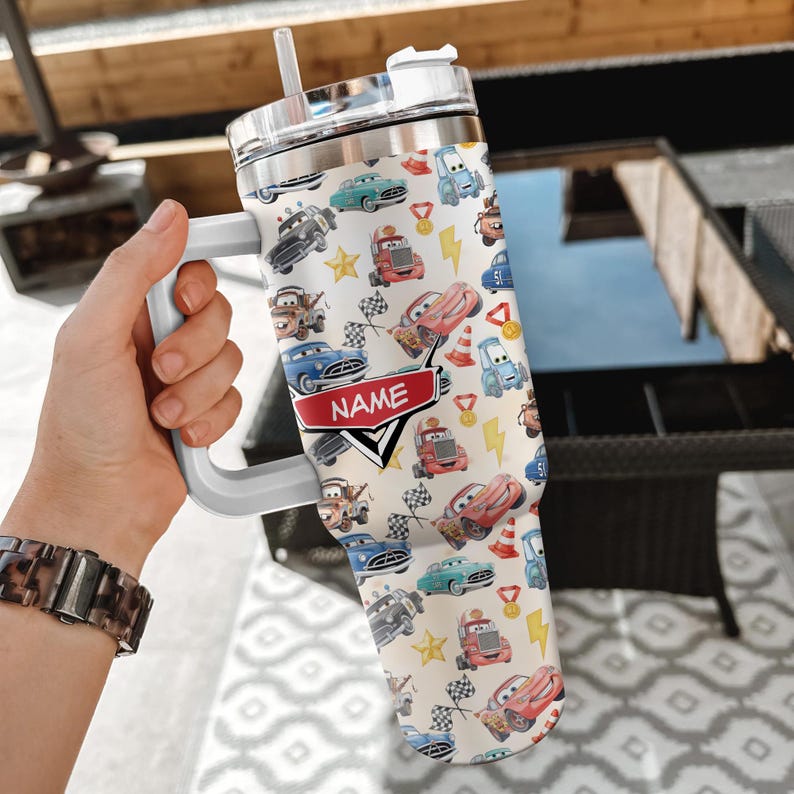 May include: A white tumbler with a handle, featuring a colorful pattern of cartoon cars, stars, and racing flags. The tumbler has a silver lid and a white straw. The word "NAME" is displayed on a red banner.