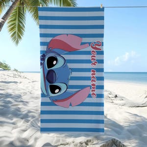 May include: Blue and white striped beach towel featuring a cartoon character with large eyes and pink ears. The text "Your name" is printed vertically in red. The towel is hanging on a clothesline on a sunny beach.