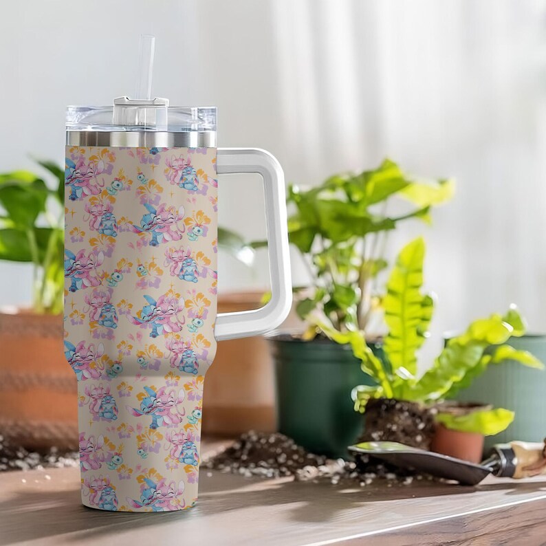 Puede incluir: Un vaso grande y aislado con asa y pajita. El vaso presenta un patr&oacute;n repetido de personajes de dibujos animados en tonos azules, rosas y amarillos sobre un fondo beige. El asa y la tapa son blancos.