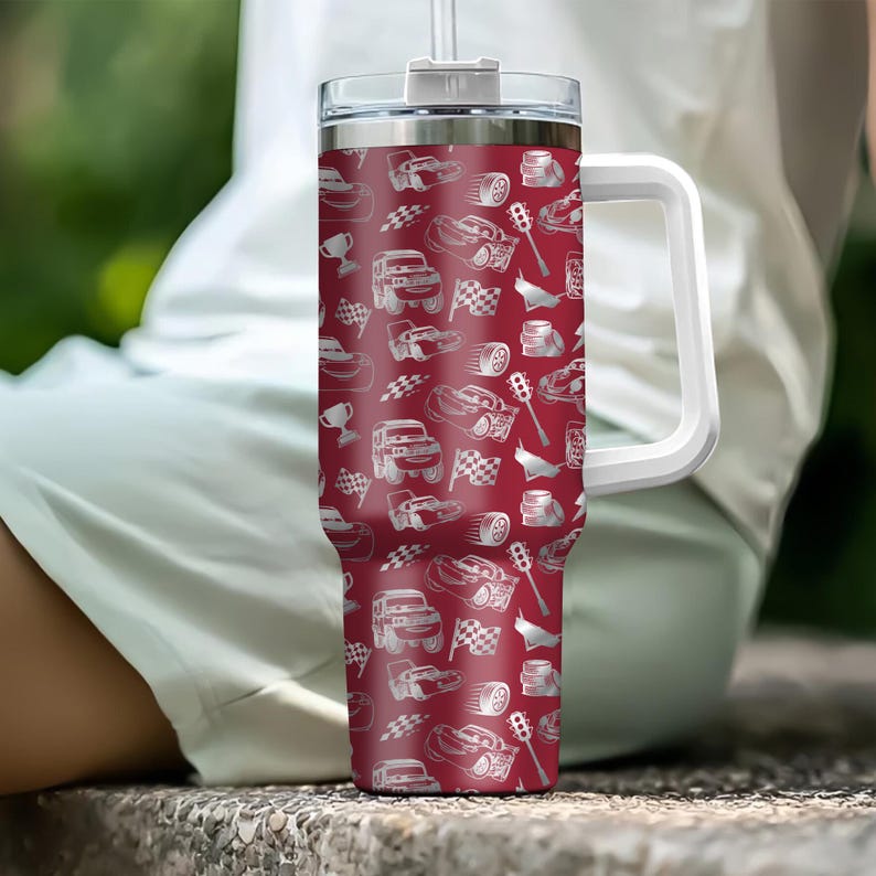 Puede incluir: Un vaso t&eacute;rmico rojo con asa y tapa blancas. El vaso est&aacute; cubierto con un estampado de coches de carreras blancos, banderas a cuadros y neum&aacute;ticos. Tiene una pajita transparente.
