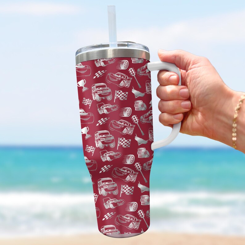 Puede incluir: Un vaso rojo con asa y pajita blancas, con un estampado de coches de carreras, banderas a cuadros y trofeos. El vaso de acero inoxidable se sostiene contra un fondo de playa.