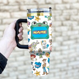 May include: A white stainless steel tumbler with a black handle and a clear lid, featuring a colorful pattern of various cartoon characters and yellow sheriff stars. A central green frog-shaped frame contains the text NAME in bold orange letters.