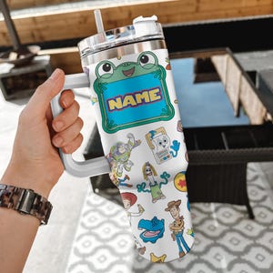 May include: A white stainless steel tumbler with a handle, a clear lid, and a straw. The surface features a colorful pattern of cartoon characters including a frog with a blue name tag, Buzz Lightyear, Woody, Jessie, and Forky on a white background.