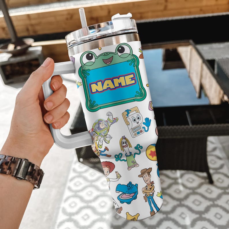 May include: A white stainless steel tumbler with a handle, a clear lid, and a straw. The surface features a colorful pattern of cartoon characters including a frog with a blue name tag, Buzz Lightyear, Woody, Jessie, and Forky on a white background.