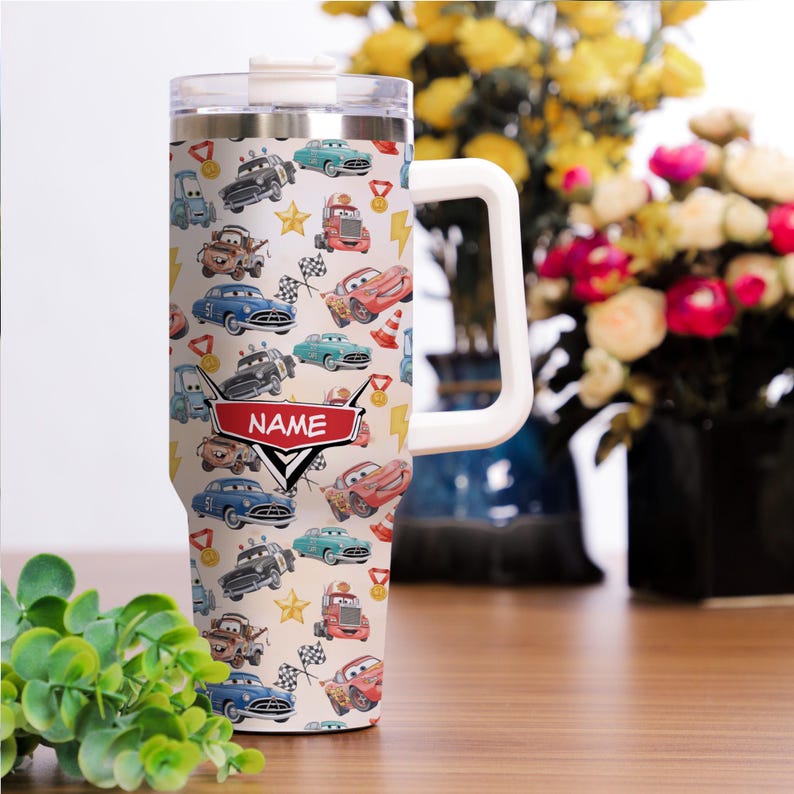 May include: A stainless steel tumbler with a white handle and lid. The tumbler is covered in a pattern of cartoon cars, stars, and racing flags. A red banner with the word "NAME" is displayed on the front.