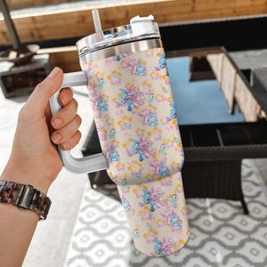Puede incluir: Un vaso de acero inoxidable con asa y pajita, con un estampado repetido de personajes de dibujos animados azules, rosas y amarillos y elementos florales sobre un fondo beige. El vaso tiene un asa y una tapa blancas.
