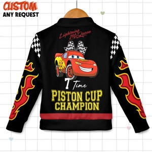 May include: Black zip-up jacket featuring Lightning McQueen from the Cars movie. The jacket has red and yellow flame designs on the sleeves, checkered flag accents, and the text "7 Time Piston Cup Champion."