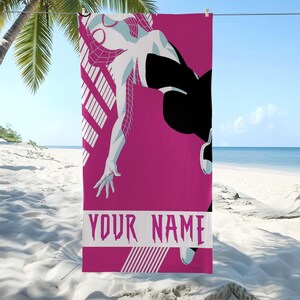 May include: A pink beach towel featuring a stylized illustration of a superhero in a dynamic pose. The character is depicted in white and black, with a web-like design. The towel includes the text "YOUR NAME" in a bold, stylized font.