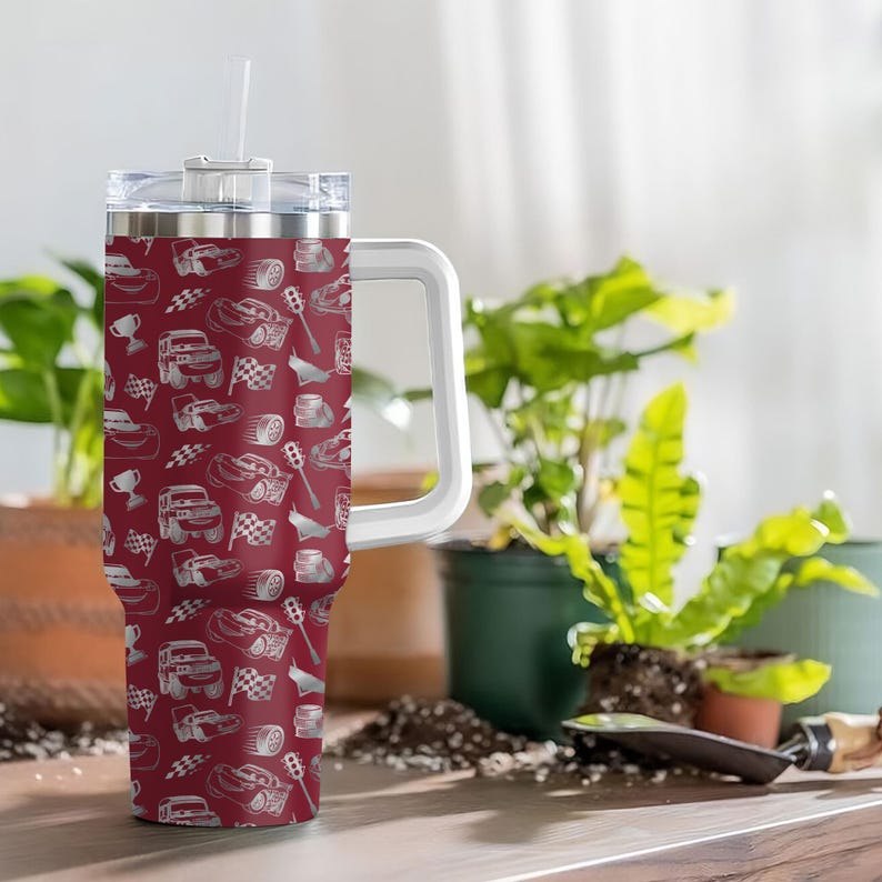 Puede incluir: Un vaso de viaje granate con asa blanca y tapa transparente. El vaso presenta un estampado de coches de carreras blancos, banderas a cuadros y trofeos. Incluye una pajita blanca.