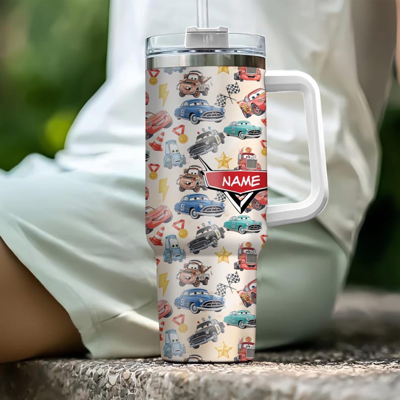 May include: A white handled tumbler with a clear lid and straw, covered in a pattern of cartoon cars, lightning bolts, and racing flags. A red banner with the word "NAME" is visible on the tumbler.