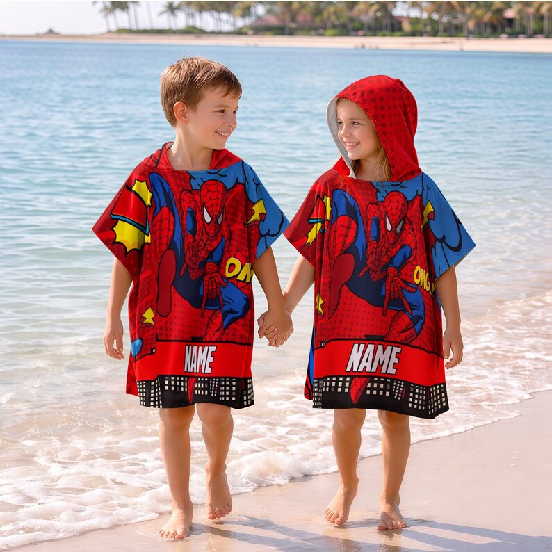 May include: Two children wearing red hooded beach ponchos with a Spider-Man graphic, holding hands on a sandy beach. The ponchos have a red base with blue and yellow accents, and the word "NAME" is printed on the bottom.