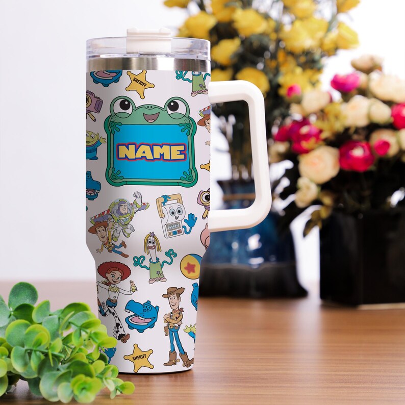 May include: A white stainless steel tumbler with a handle and a clear lid, featuring a repeating pattern of cartoon characters including a cowboy, a space ranger, a cowgirl, and a frog-shaped frame with the text NAME printed in yellow.
