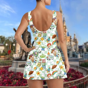 Puede incluir: Vestido sin mangas con un estampado de cuadros blancos y verde claro, adornado con personajes y objetos de dibujos animados coloridos. El vestido tiene la espalda escotada y una falda acampanada. Los personajes de dibujos animados incluyen un vaquero, un astronauta y un extraterrestre.