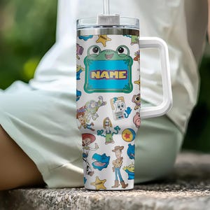 May include: A white stainless steel tumbler with a handle and straw, featuring a colorful pattern of various animated toy characters, yellow sheriff stars, and a central green-bordered rectangular frame with the word NAME printed in orange.
