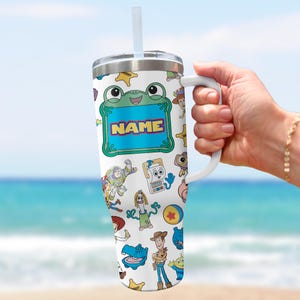 May include: A white stainless steel tumbler with a handle, a clear lid, and a straw. The surface features a colorful pattern of various cartoon characters and a central green frog-shaped frame with the word NAME printed in yellow and blue letters.