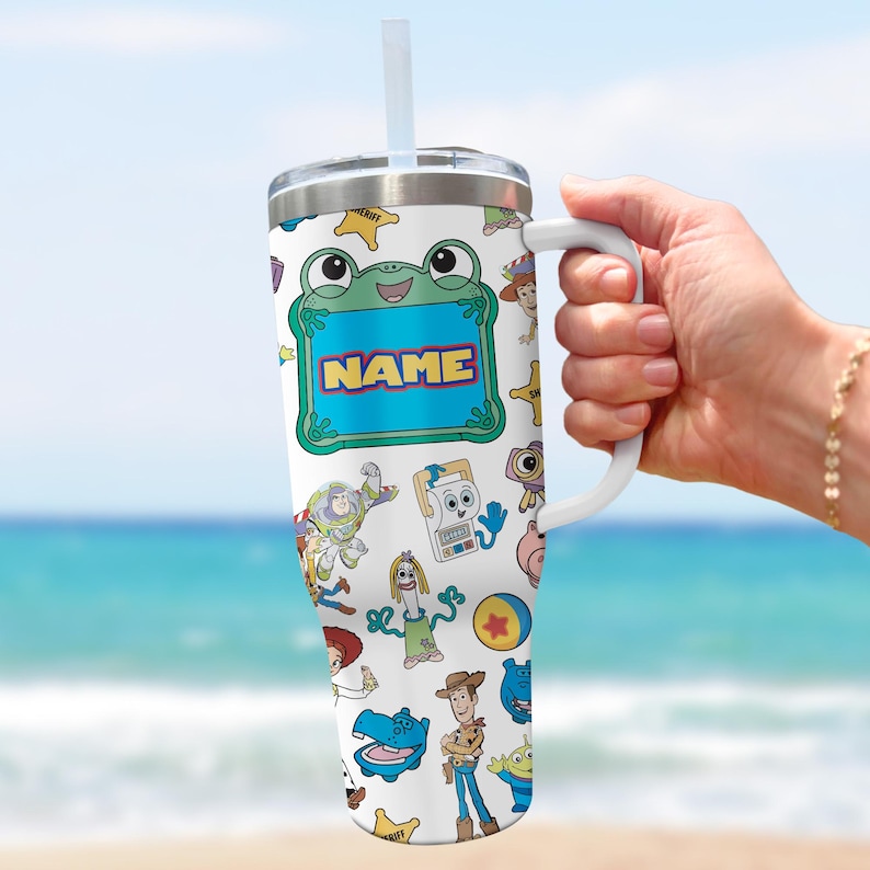 May include: A white stainless steel tumbler with a handle, a clear lid, and a straw. The surface features a colorful pattern of various cartoon characters and a central green frog-shaped frame with the word NAME printed in yellow and blue letters.