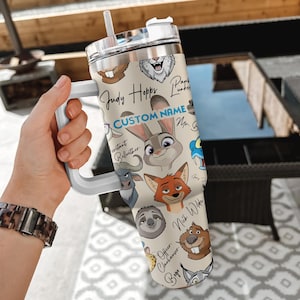 Puede incluir: Un vaso de acero inoxidable con personajes de dibujos animados de Zootopia. El vaso tiene un asa blanca y una tapa transparente con pajita. El dise&ntilde;o incluye a Judy Hopps, Nick Wilde y otros personajes, con espacio para un nombre personalizado.