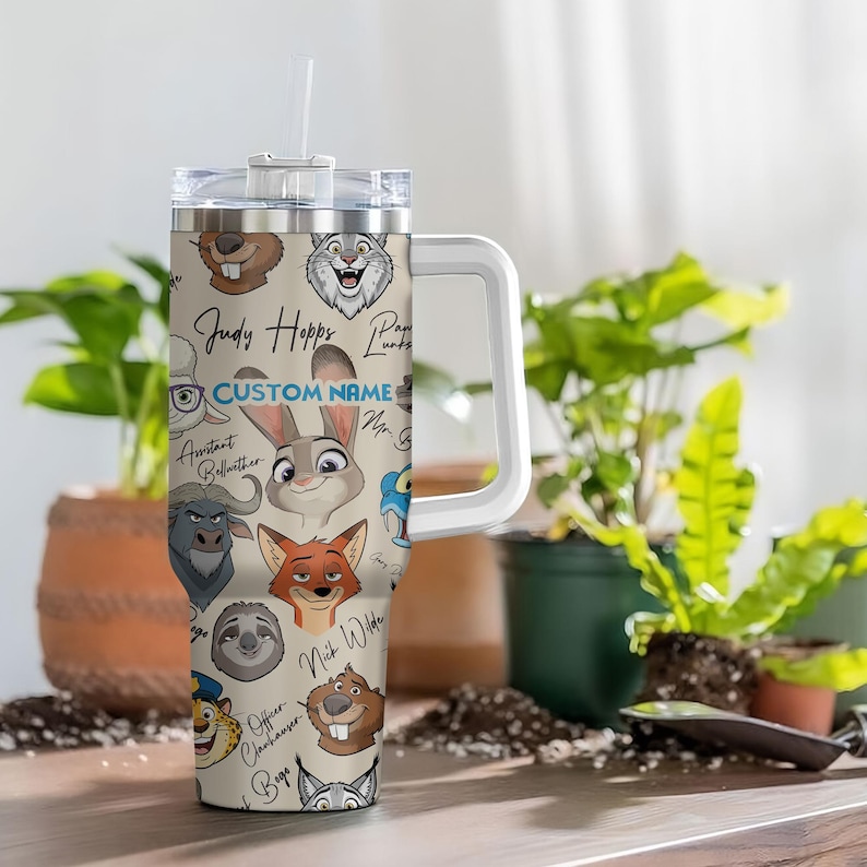 Puede incluir: Un vaso con asa blanca con personajes de dibujos animados de Zootopia. El vaso tiene una tapa transparente y una pajita blanca. El vaso tiene las palabras "Judy Hopps" y "Custom Name" impresas. El vaso est&aacute; rodeado de plantas verdes.