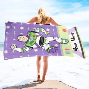 Puede incluir: Una toalla de playa morada con una ilustraci&oacute;n de dibujos animados de Buzz Lightyear de Toy Story. La toalla tiene un patr&oacute;n repetido de la cara de Buzz Lightyear y cohetes. El lado derecho de la toalla tiene el texto "Your Name".