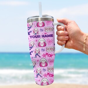 Pode incluir: Um copo rosa com uma al&ccedil;a e canudo brancos, com um padr&atilde;o repetido de um personagem de super-her&oacute;i, teias de aranha e a palavra "POW!". O texto "YOUR NAME" tamb&eacute;m &eacute; vis&iacute;vel. O copo &eacute; segurado por uma pessoa.