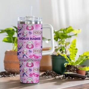Pode incluir: Um copo personalizado com um padr&atilde;o repetido de um personagem de super-her&oacute;i em rosa, roxo e branco. O copo tem uma al&ccedil;a branca, uma tampa transparente e um canudo. O texto "YOUR NAME" est&aacute; impresso no copo.