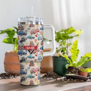 May include: A personalized tumbler with a white handle and straw. The tumbler features a repeating pattern of cartoon cars in various colors, including red, blue, and green, on a cream background. A red banner with the word "NAME" is also visible.