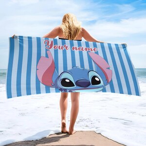 May include: A blue and white striped beach towel featuring a cartoon character with large black eyes and pink ears. The towel has the text "Your name" in a pink script font. The towel is held up by a person on a beach.