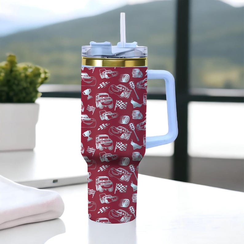 Puede incluir: Un vaso grande y aislado con un fondo rojo y un patr&oacute;n de coches de carreras blancos. El vaso tiene un asa y una tapa azul claro, un borde dorado y una pajita blanca. Est&aacute; dise&ntilde;ado para contener una gran cantidad de l&iacute;quido.