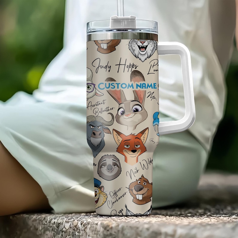 Puede incluir: Un vaso beige con asa y tapa blancas, con ilustraciones de personajes de dibujos animados. El vaso tiene el texto "CUSTOM NAME" en azul y nombres de personajes como "Judy Hopps" y "Nick Wilde".