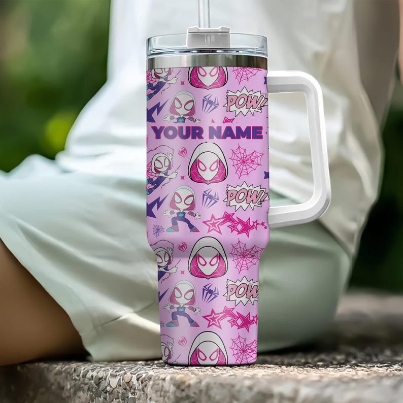 Pode incluir: Um copo rosa com uma al&ccedil;a e tampa brancas, com um padr&atilde;o de ilustra&ccedil;&otilde;es da Spider-Gwen, teias de aranha e texto "POW!". O texto "YOUR NAME" est&aacute; impresso em roxo. O copo tem uma palhinha transparente.