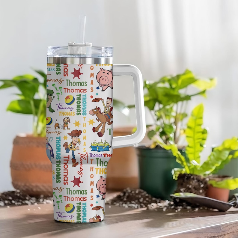 Pu&ograve; includere: Un bicchiere termico bianco in acciaio inossidabile con manico e coperchio trasparente, caratterizzato da un motivo ripetuto di personaggi di Toy Story come Woody, Bullseye, Hamm e gli alieni, con il nome Thomas e stelle colorate.