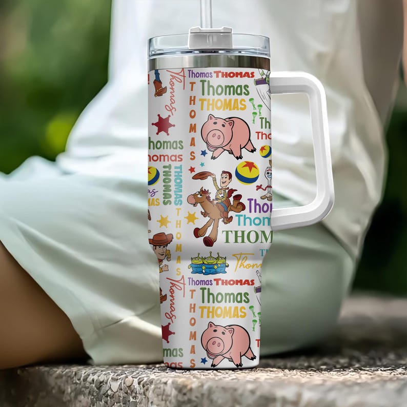 Pu&ograve; includere: Un bicchiere termico bianco in acciaio inossidabile con manico e coperchio, caratterizzato da un motivo ripetuto di personaggi di Toy Story come Woody, Hamm e gli Alieni, intervallati da stelle, palline e il nome Thomas in vari font e colori.