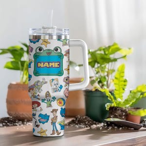 May include: A white stainless steel tumbler with a handle and clear lid featuring a colorful pattern of various cartoon characters, including a cowboy, a space ranger, and a frog-shaped frame with the word NAME printed in the center.
