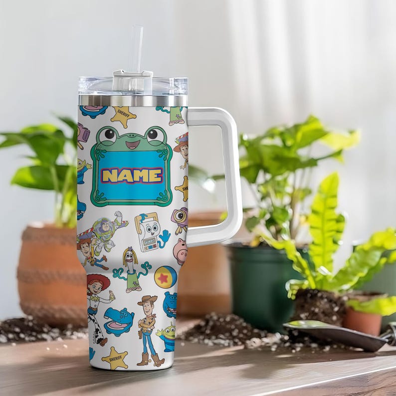 May include: A white stainless steel tumbler with a handle and clear lid featuring a colorful pattern of various cartoon characters, including a cowboy, a space ranger, and a frog-shaped frame with the word NAME printed in the center.