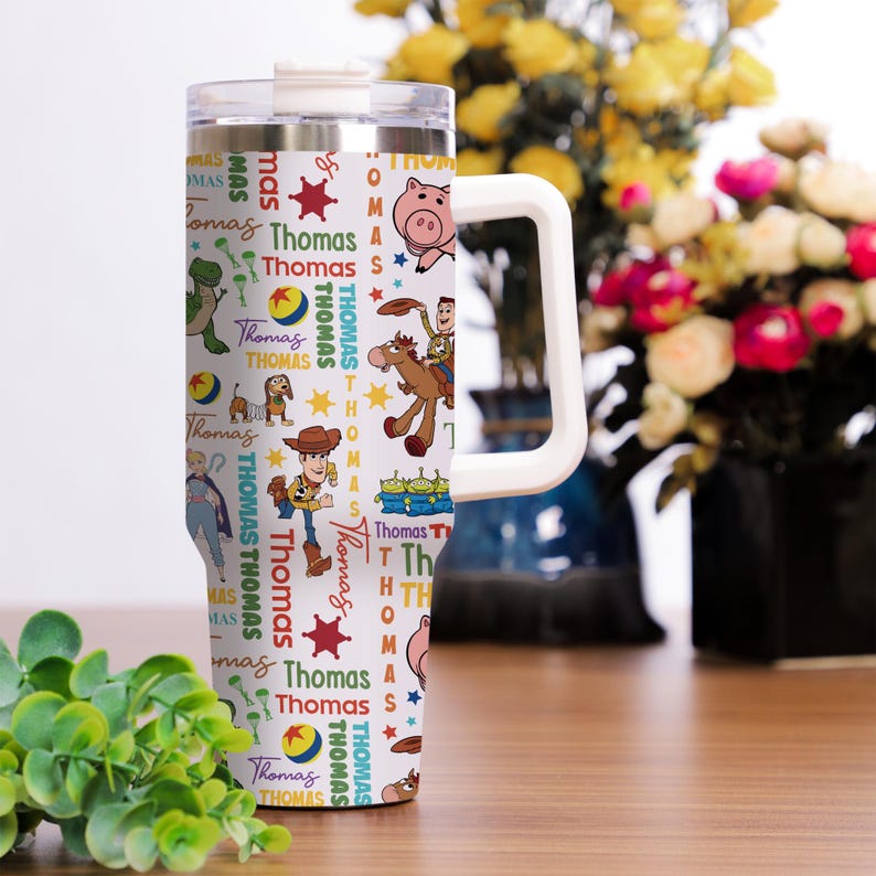 Pu&ograve; includere: Un bicchiere termico bianco in acciaio inossidabile con manico e coperchio trasparente, caratterizzato da un motivo ripetuto di personaggi di Toy Story come Woody, Rex e Hamm, intervallati dal nome Thomas in vari colori e piccole stelle.
