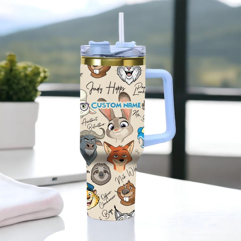 Puede incluir: Un vaso t&eacute;rmico beige con personajes de dibujos animados de Zootopia, con un asa y tapa azul claro. El vaso tiene una pajita y detalles dorados. El texto "CUSTOM NAME" es visible.
