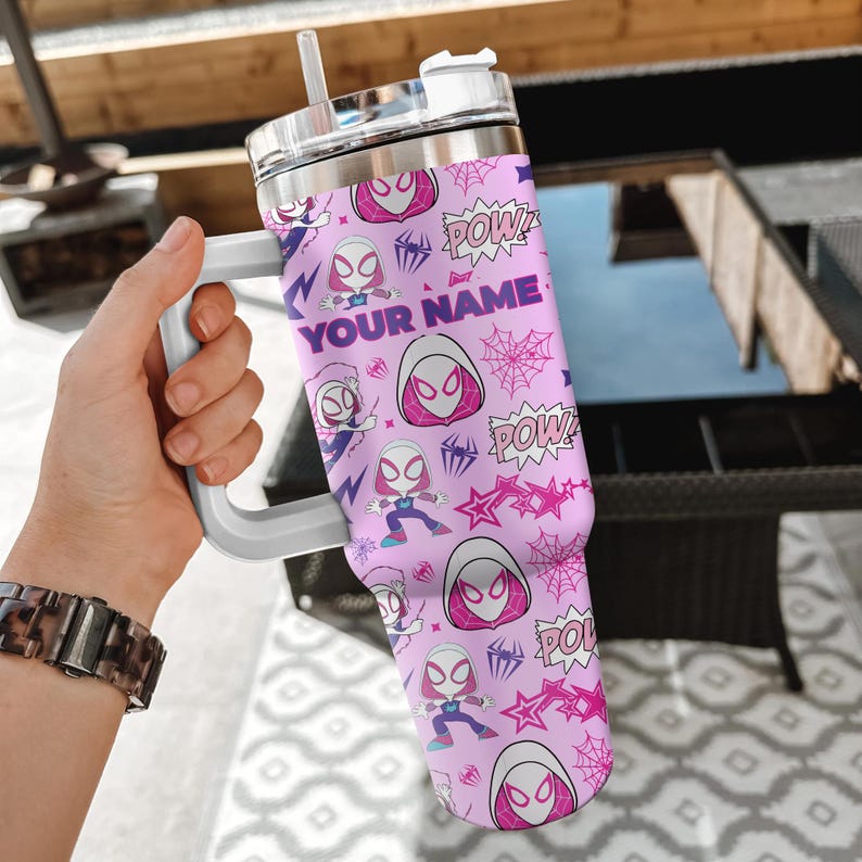 Pode incluir: Um copo rosa com uma al&ccedil;a e tampa brancas, apresentando um padr&atilde;o repetido de um personagem de super-her&oacute;i, teias de aranha e a palavra "POW!" em estilo de hist&oacute;ria em quadrinhos. O texto "YOUR NAME" tamb&eacute;m &eacute; vis&iacute;vel.